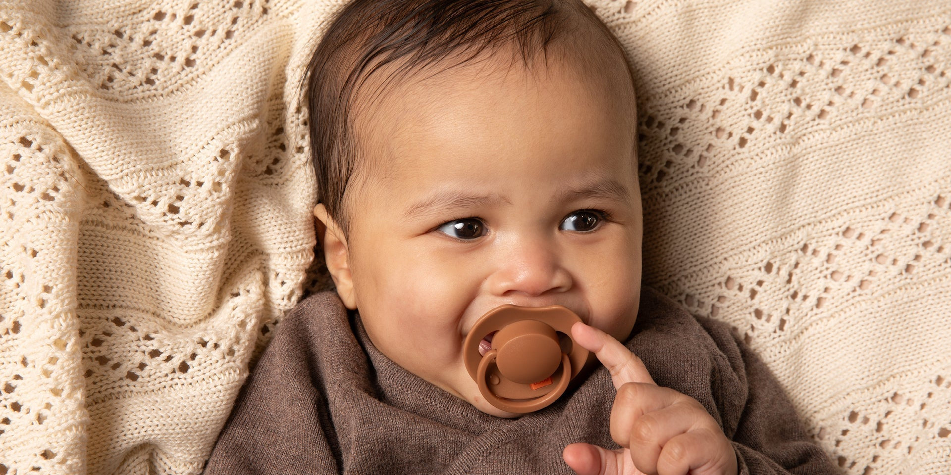Un bébé de 6 mois est allongé sur une couverture beige. Le bébé sourit et a une sucette Difrax LOVI de la collection Prime, de couleur marron.