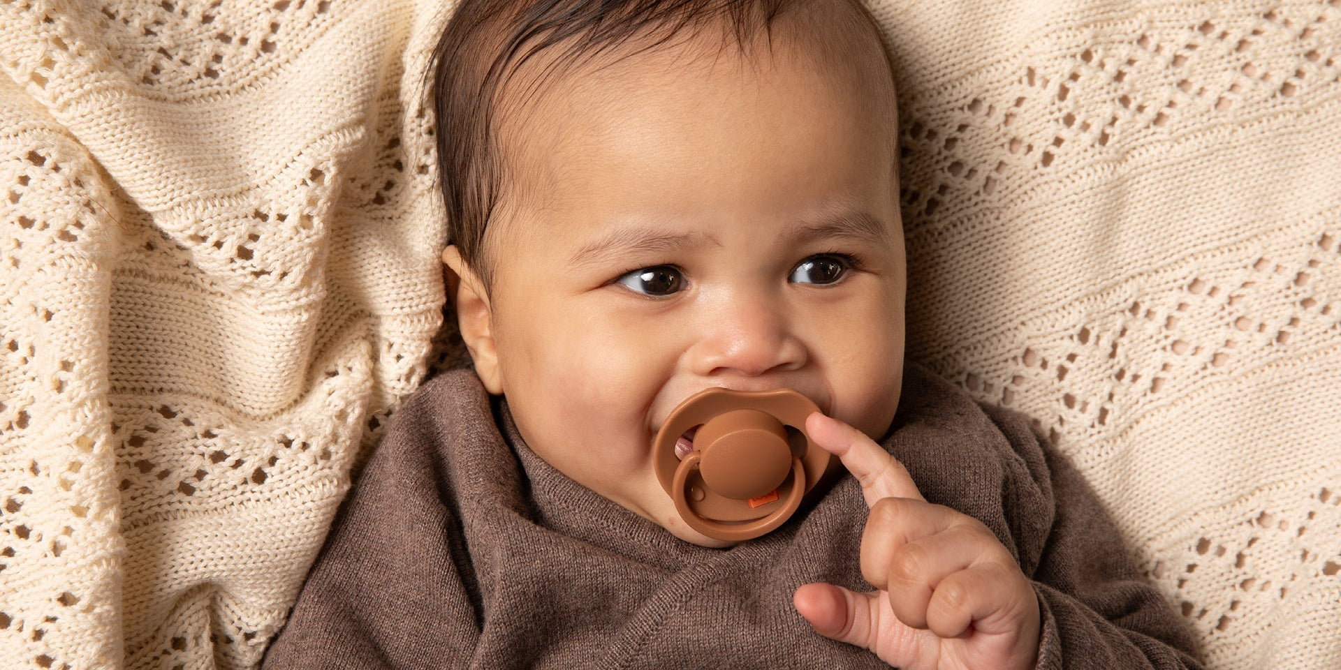 Un bébé de 6 mois est allongé sur une couverture beige. Le bébé sourit et a une sucette Difrax LOVI de la collection Prime, de couleur marron.