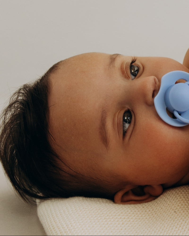 Bébé allongé tenant une sucette bleue Prime dans ses mains et portant une autre sucette bleue en bouche.