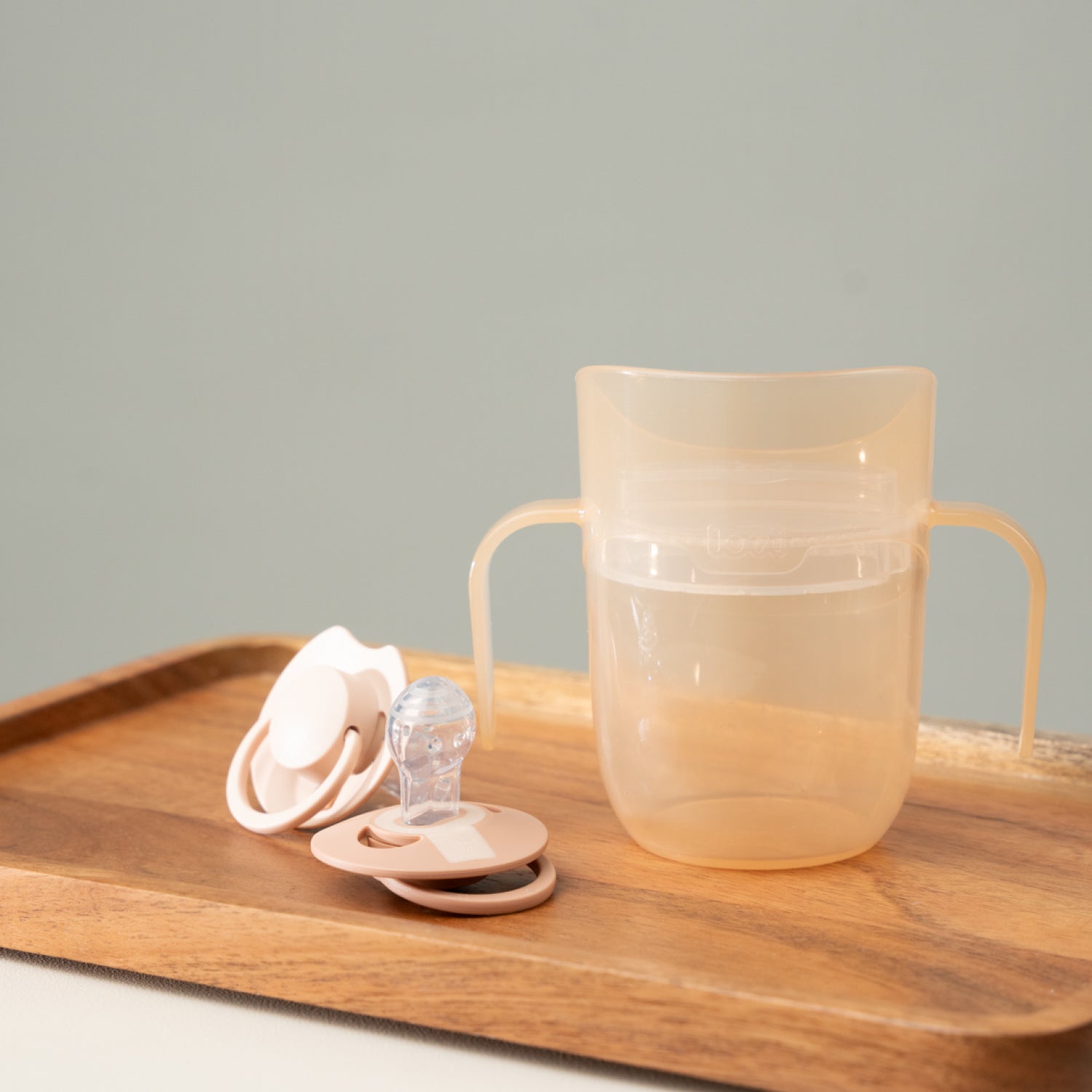 Gobelet d’apprentissage Difrax LOVI Genius couleur nude sur une table, à côté de deux sucettes dynamiques – produit bébé élégant et fonctionnel