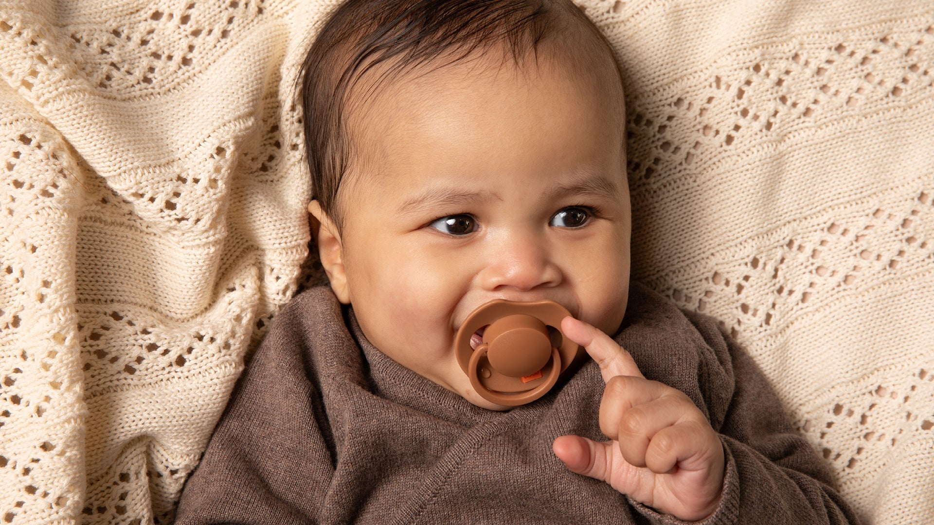 Un bébé de 6 mois est allongé sur une couverture beige. Le bébé sourit et a une sucette Difrax LOVI de la collection Prime, de couleur marron.