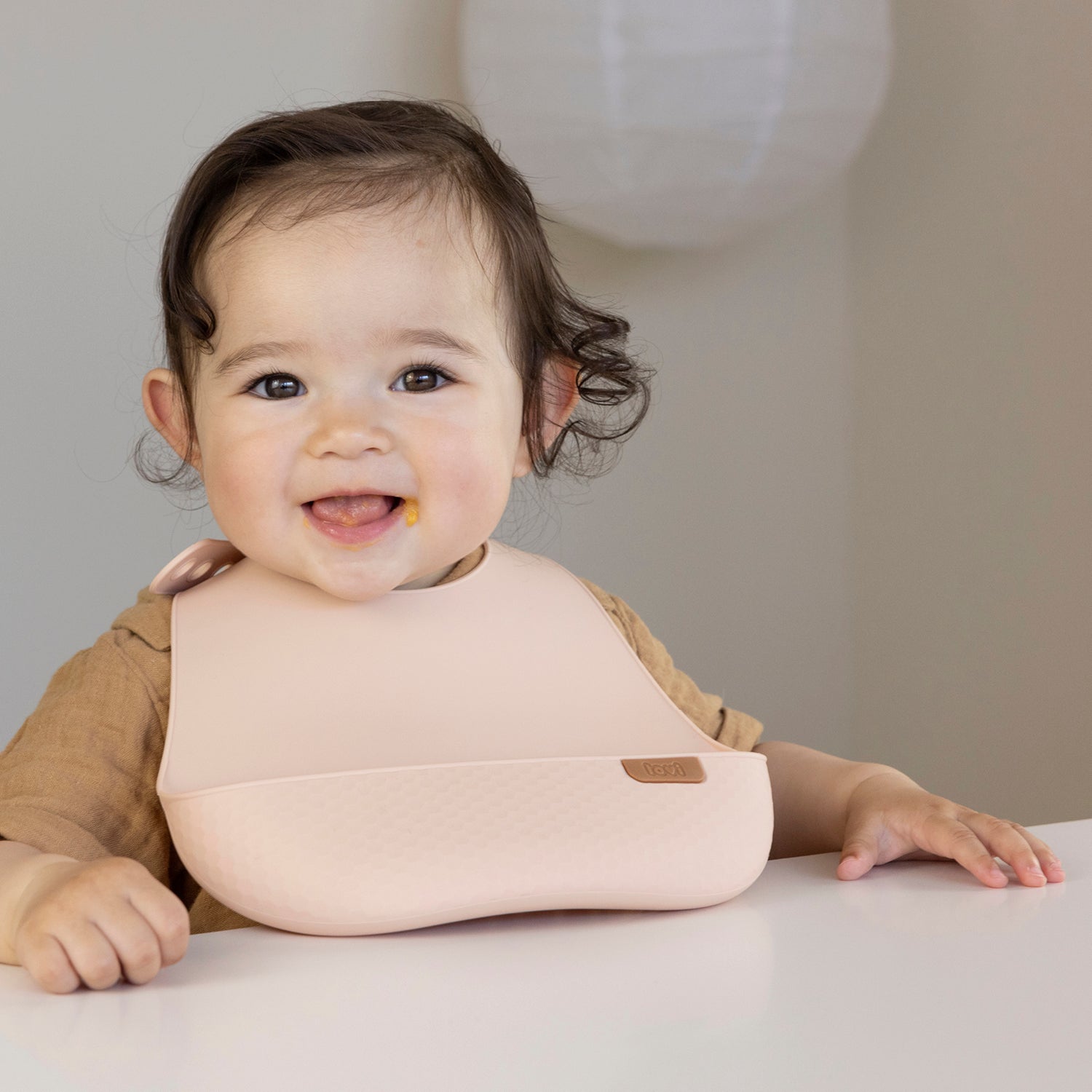 Un bambin heureux est assis dans la chaise de bébé et déguste son repas avec la plaque en silicone couleur nude.