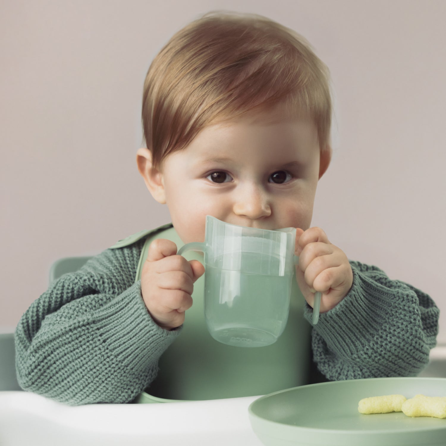 Un tout-petit est assis à la table à manger et tient le gobelet d’apprentissage Difrax LOVI Genius couleur pistache, prêt à boire de façon autonome